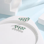 May include: A pair of silver ear climbers, each featuring a leaf design with multiple emerald-green gemstones. The earrings are presented on a white, curved surface, set against a light blue background.