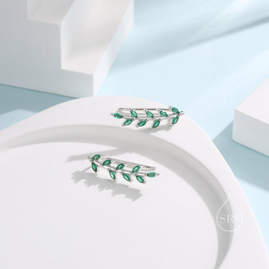 May include: A pair of silver ear climbers, each featuring a leaf design with multiple emerald-green gemstones. The earrings are presented on a white, curved surface, set against a light blue background.