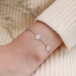 May include: A silver chain bracelet with five small white daisy charms with yellow centres. The bracelet is on a person&