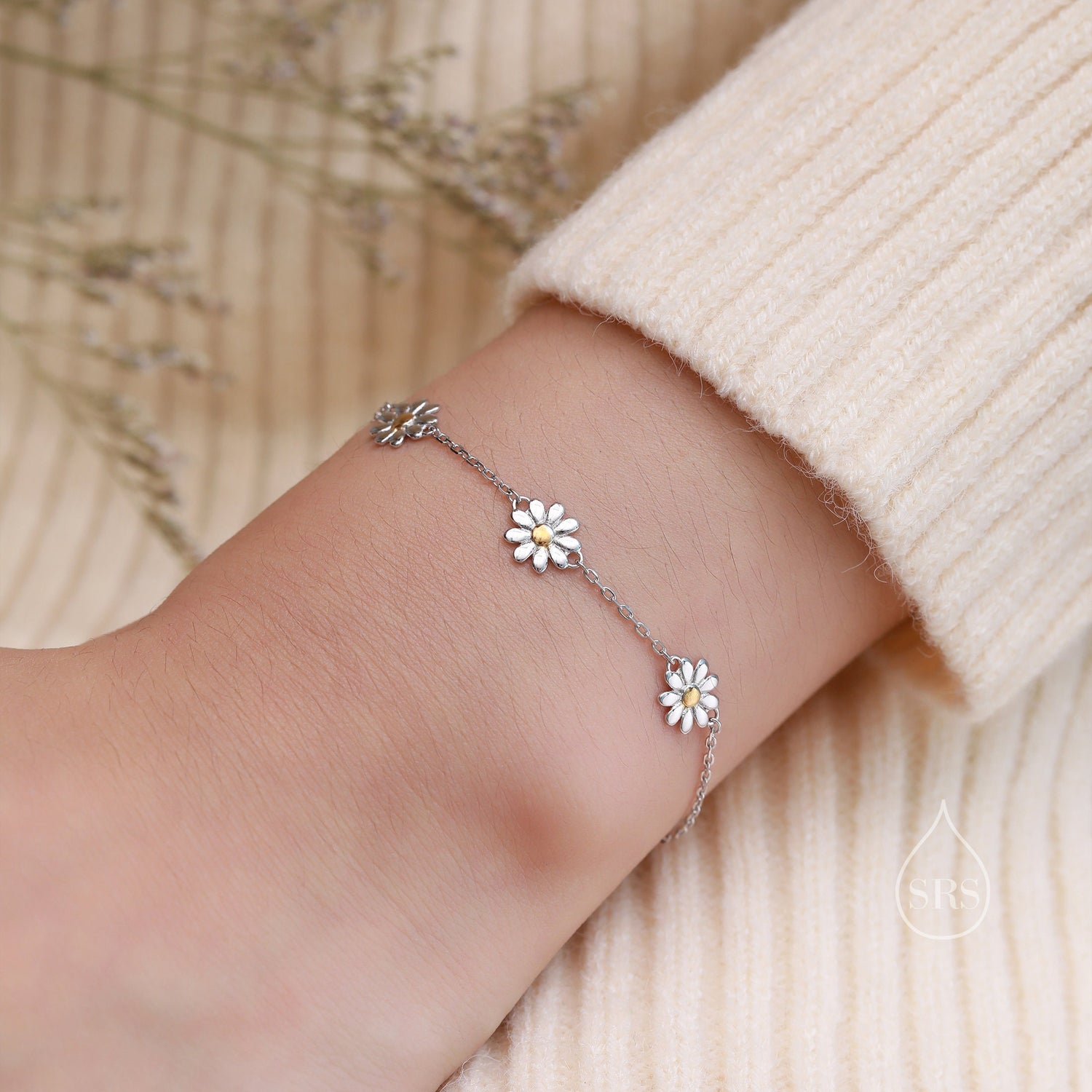May include: A silver chain bracelet with five small white daisy charms with yellow centres. The bracelet is on a person&