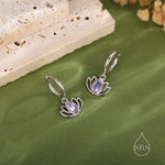 May include: Silver-coloured lotus flower earrings with a pearlescent stone centre. The earrings are displayed on a green surface with a textured background and a logo in the bottom right corner.