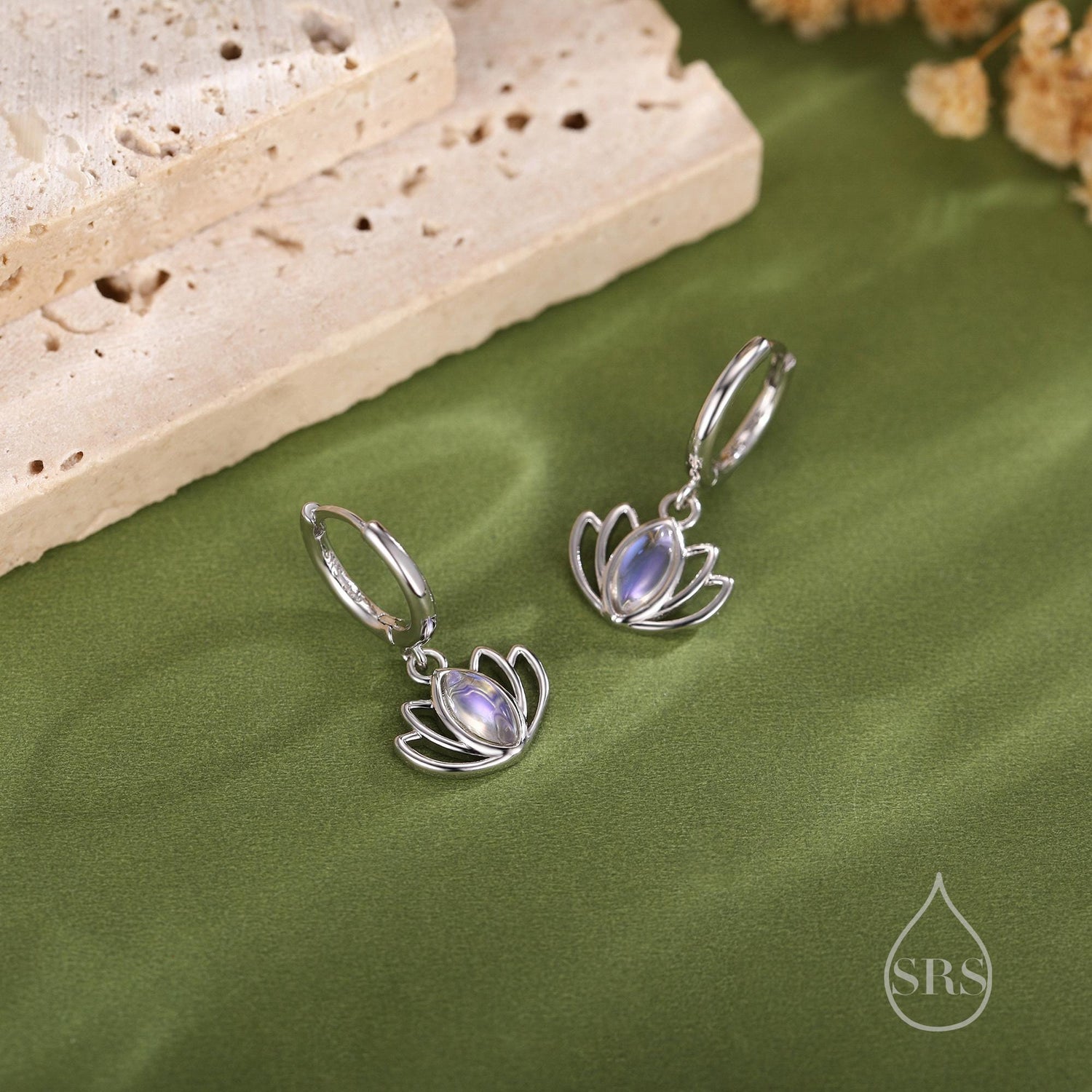 May include: Silver-coloured lotus flower earrings with a pearlescent stone centre. The earrings are displayed on a green surface with a textured background and a logo in the bottom right corner.