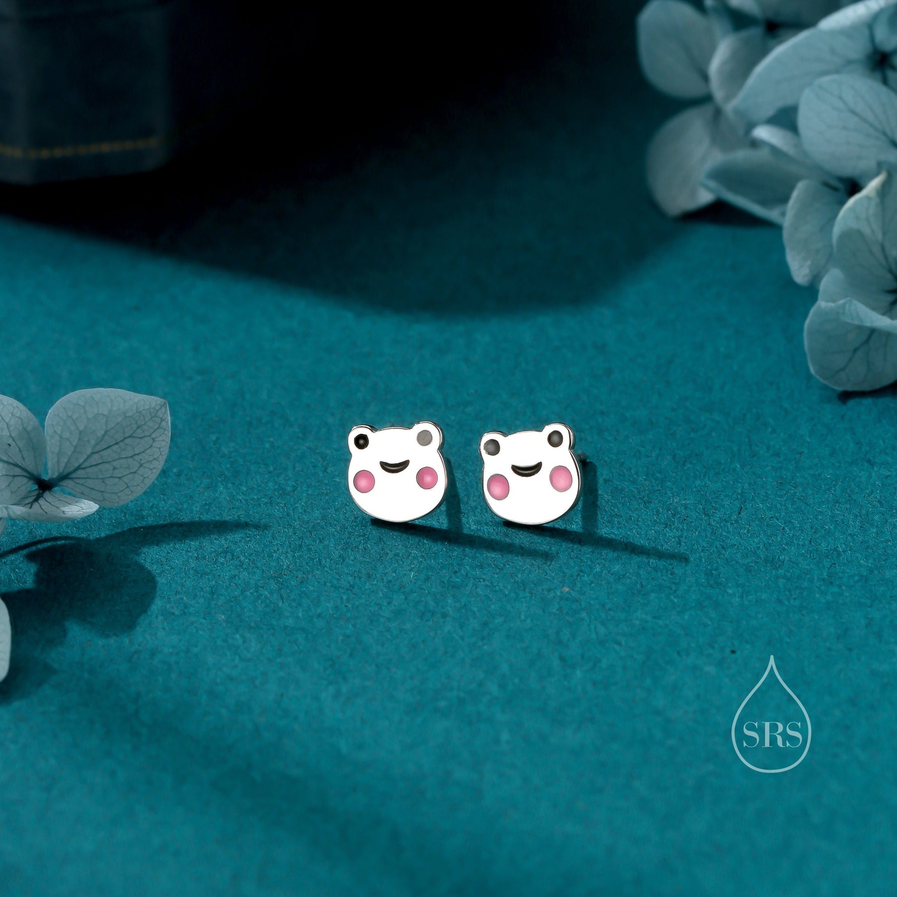 May include: A pair of silver frog stud earrings with pink accents. The frogs have a smiling face and are set against a teal background.