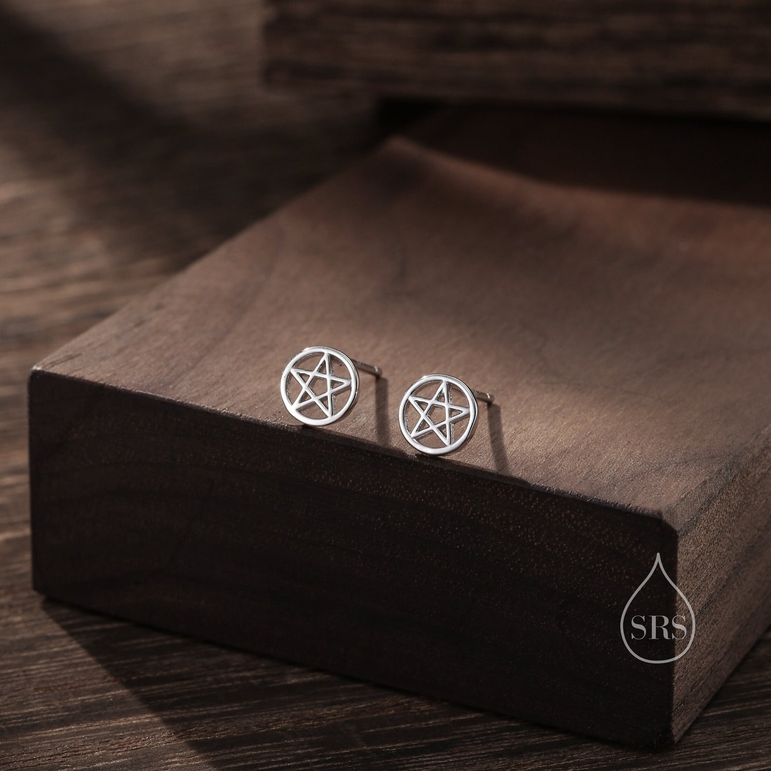 May include: A pair of silver stud earrings featuring a pentagram design. The earrings are displayed on a wooden surface.