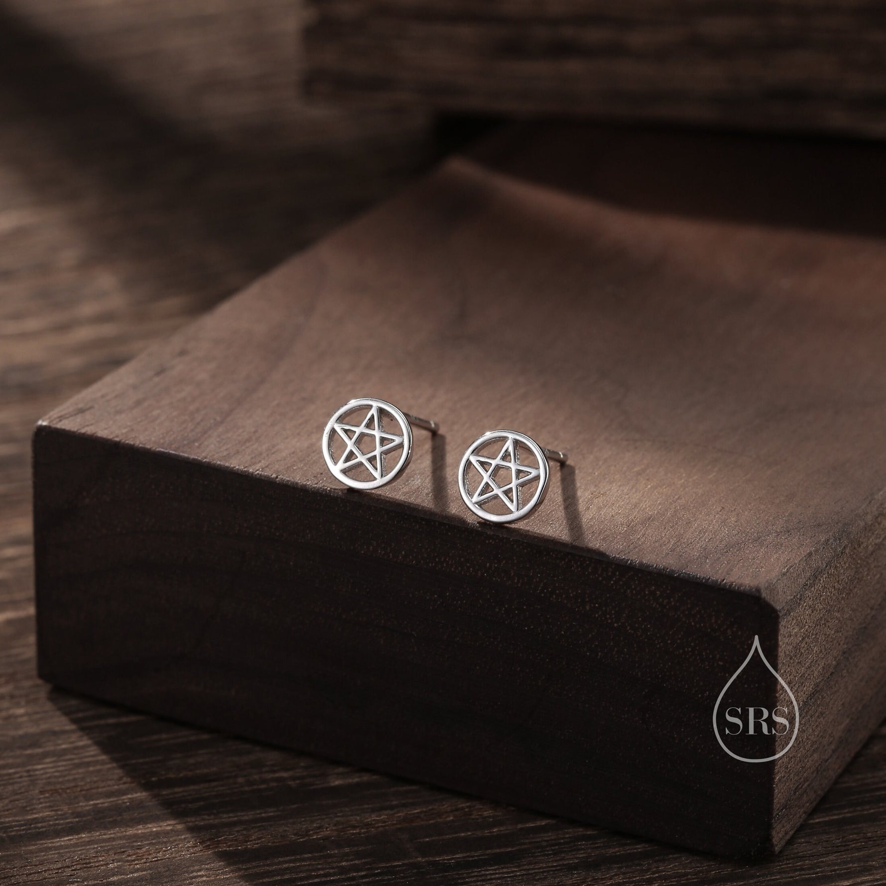 May include: A pair of silver stud earrings featuring a pentagram design. The earrings are displayed on a wooden surface.