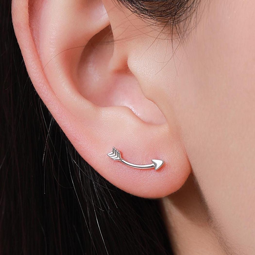 May include: A silver arrow earring with a curved design. The earring is worn in the left earlobe.