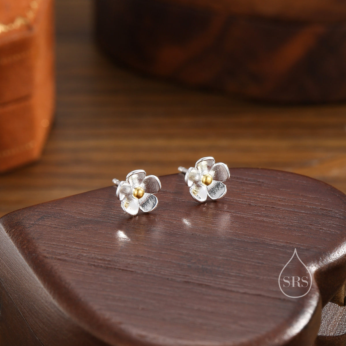 May include: Silver flower stud earrings with a gold-coloured centre. The earrings are displayed on a dark wooden surface. The floral design features five petals.
