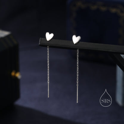May include: A pair of silver heart-shaped dangle earrings with delicate chains. The earrings are displayed on a black surface.