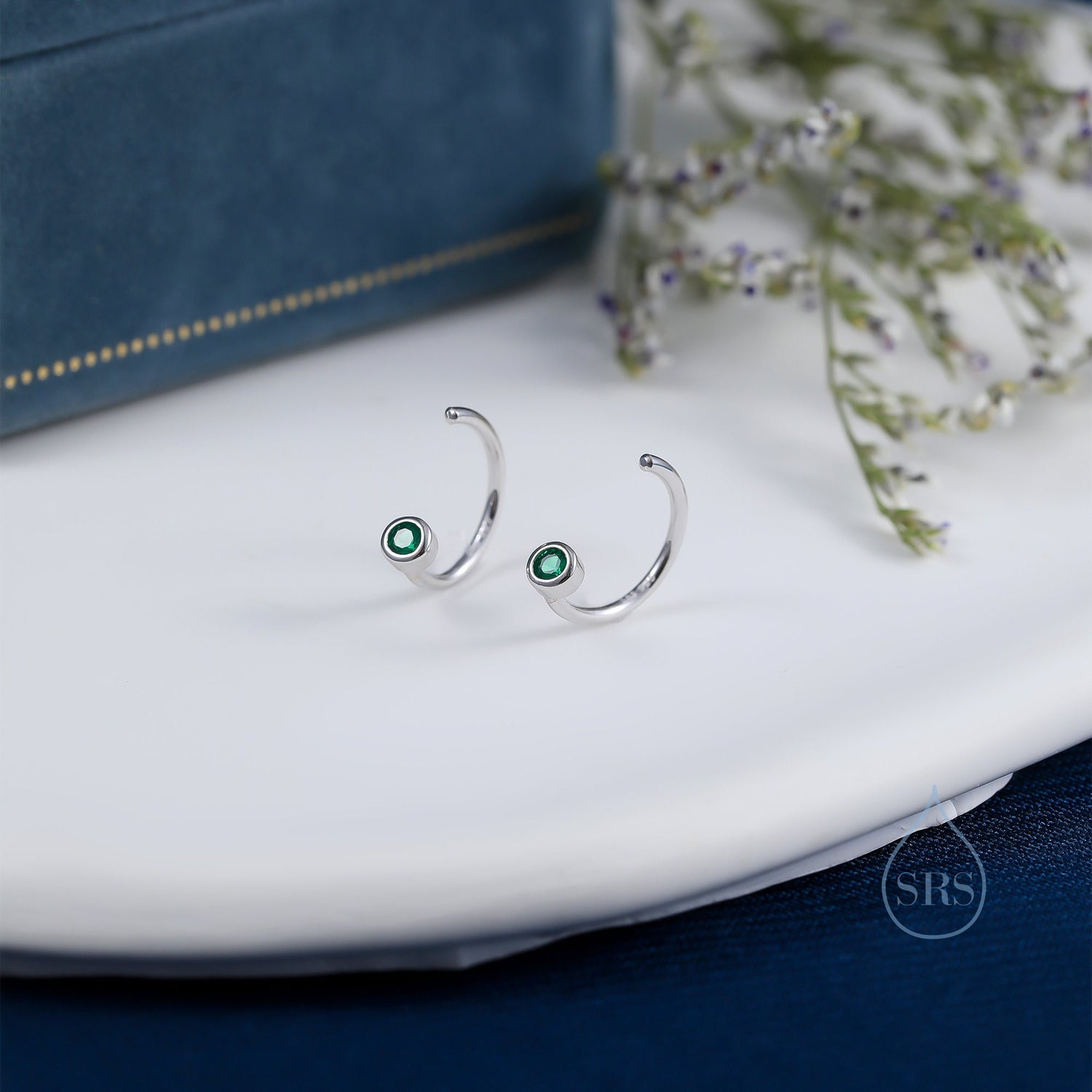 May include: Two silver hoop earrings, each with a small, round, emerald-green gemstone. The earrings have a curved design and are presented on a white surface, with a blue box in the background.