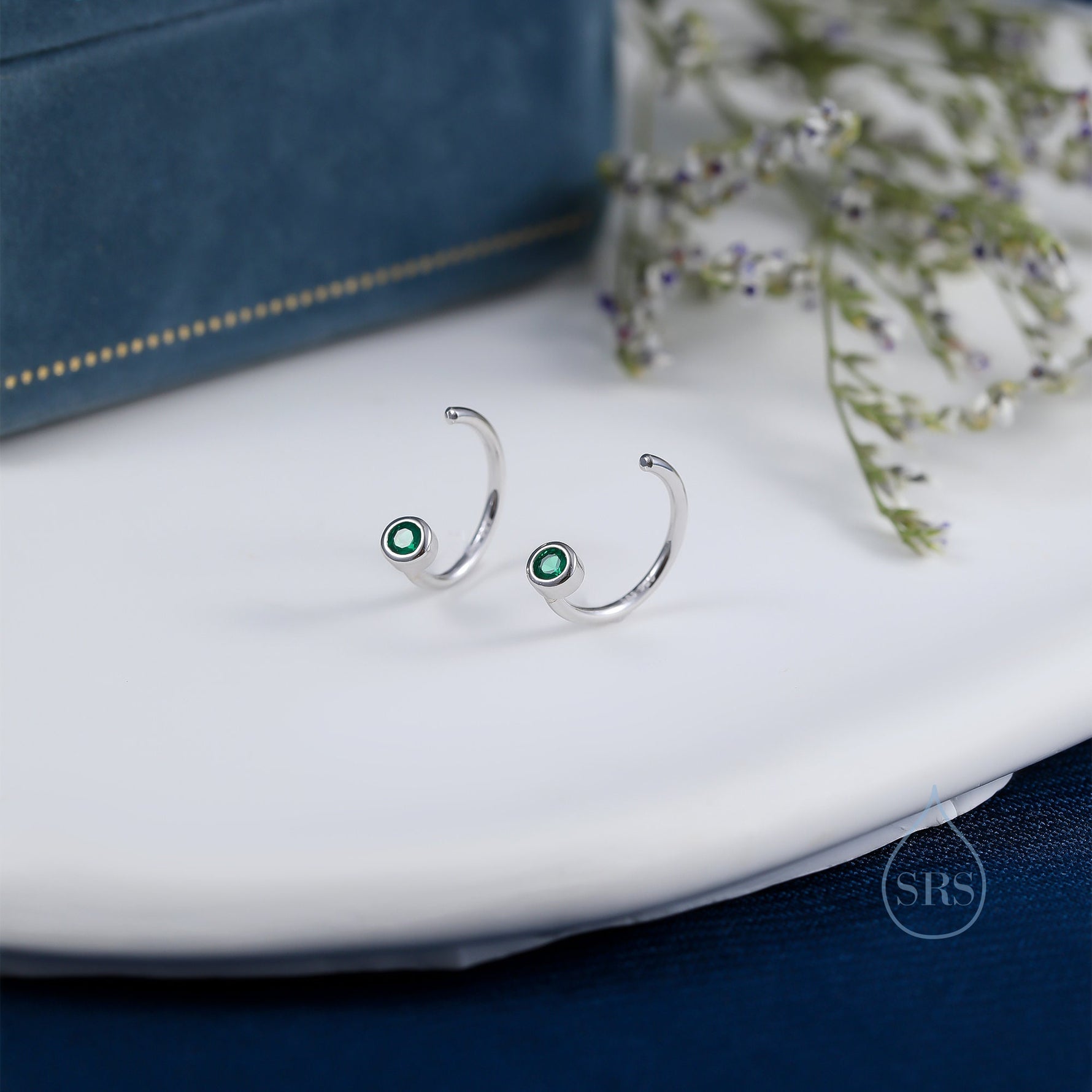 May include: Two silver hoop earrings, each with a small, round, emerald-green gemstone. The earrings have a curved design and are presented on a white surface, with a blue box in the background.