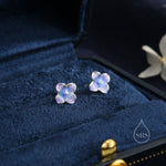 May include: A pair of silver stud earrings featuring a blue and purple flower design. The earrings are displayed in a blue velvet jewellery box.