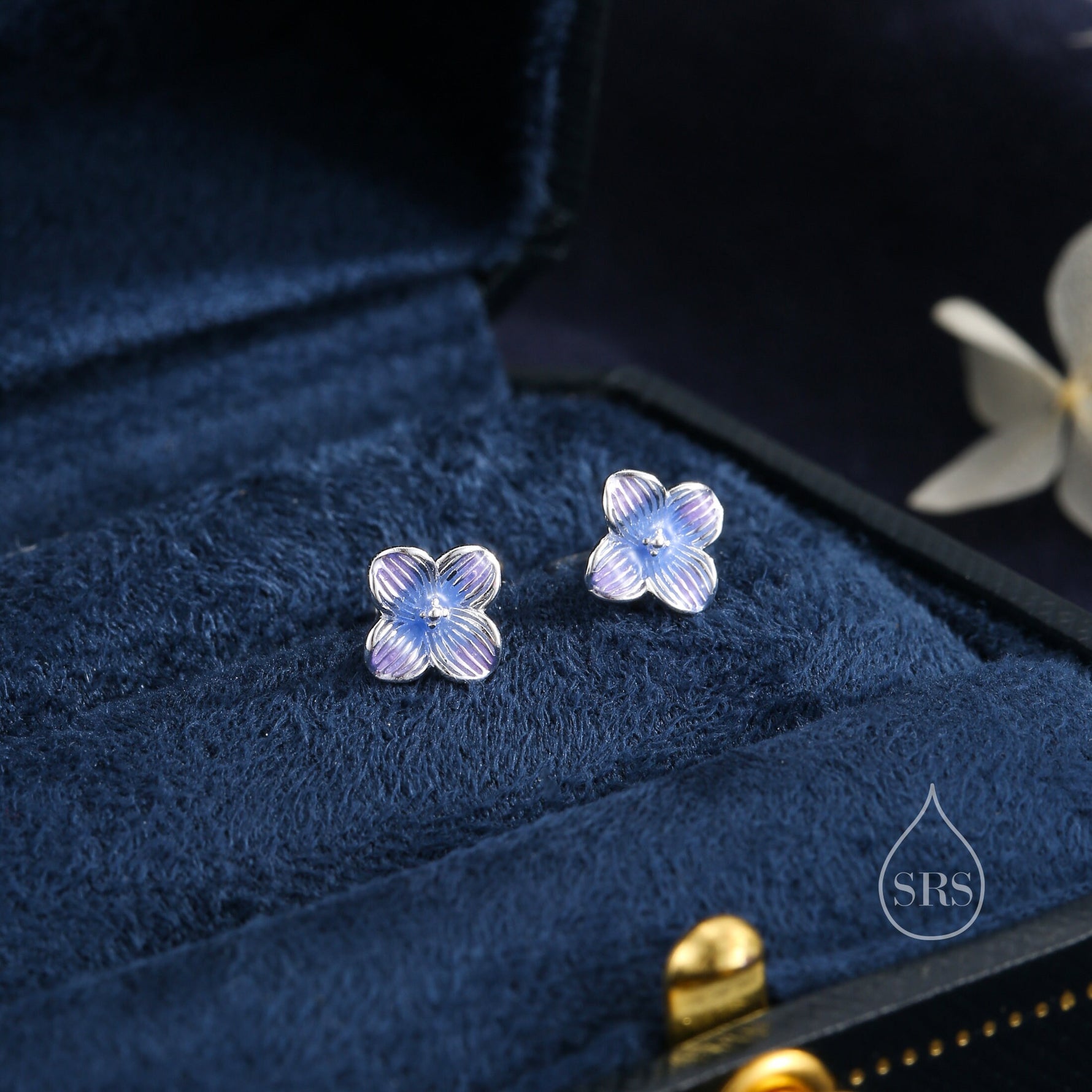 May include: A pair of silver stud earrings featuring a blue and purple flower design. The earrings are displayed in a blue velvet jewellery box.