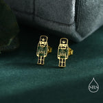 May include: A pair of gold-coloured stud earrings with a miniature nutcracker soldier design. The earrings have black and green enamel details, with a crown, uniform, and boots. The earrings are displayed on a dark green surface.