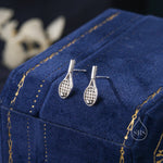 May include: A pair of silver tennis racket earrings with a small ball detail. The earrings are displayed on a blue velvet background.