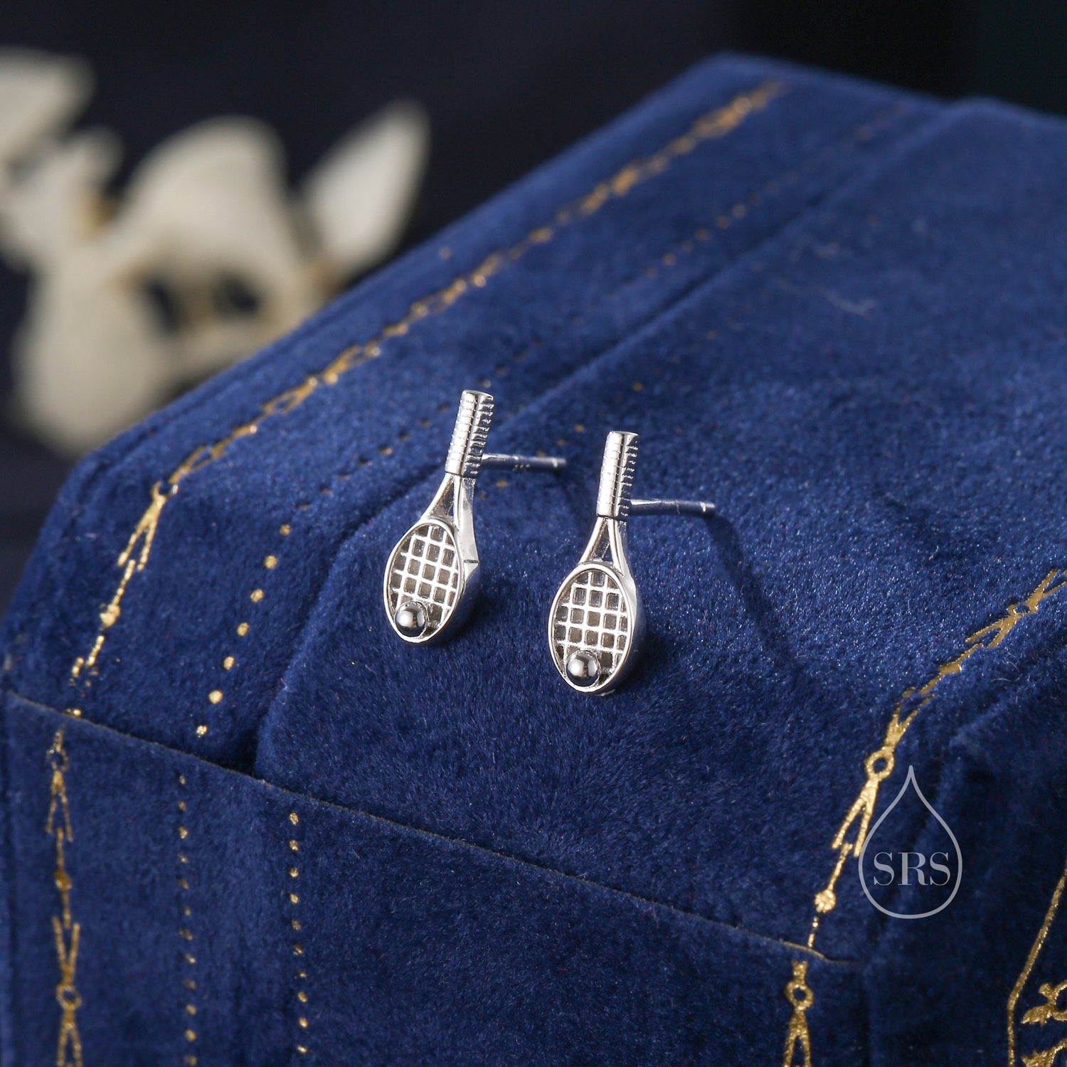 May include: A pair of silver tennis racket earrings with a small ball detail. The earrings are displayed on a blue velvet background.