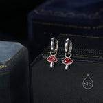 May include: A pair of silver hoop earrings with red mushroom charms. The mushrooms have white stems and white dots on the red caps.