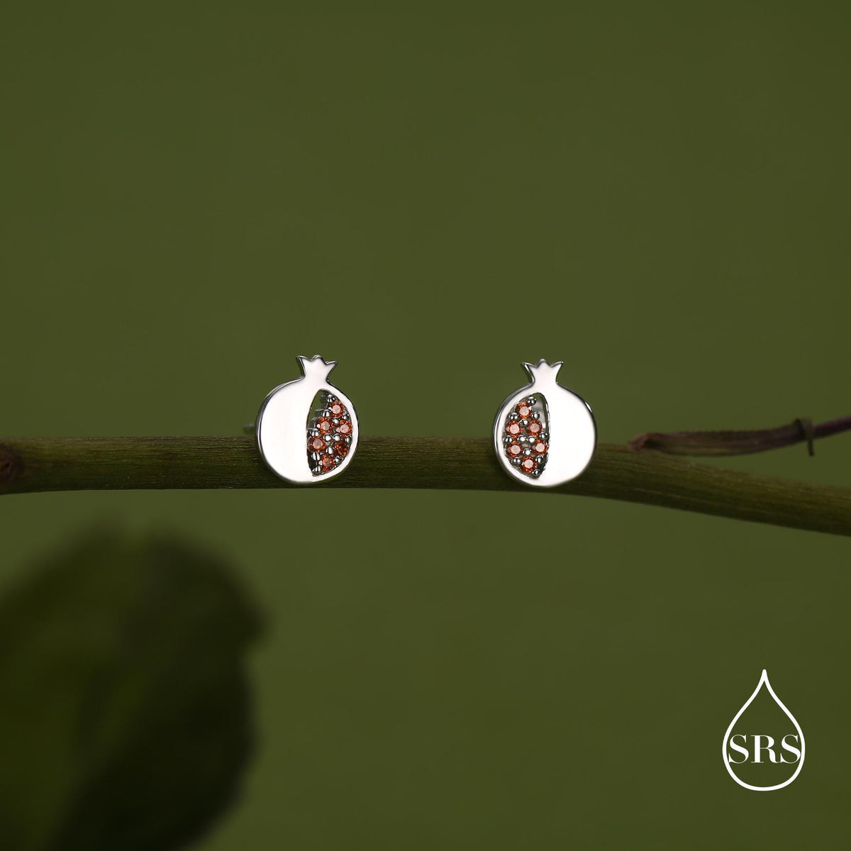 May include: A pair of silver-coloured stud earrings in the shape of pomegranates. The earrings have a white outer shell and are filled with small, reddish-brown stones. The earrings are displayed on a green branch against a green background.