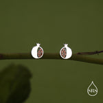 May include: A pair of silver-coloured stud earrings in the shape of pomegranates. The earrings have a white outer shell and are filled with small, reddish-brown stones. The earrings are displayed on a green branch against a green background.