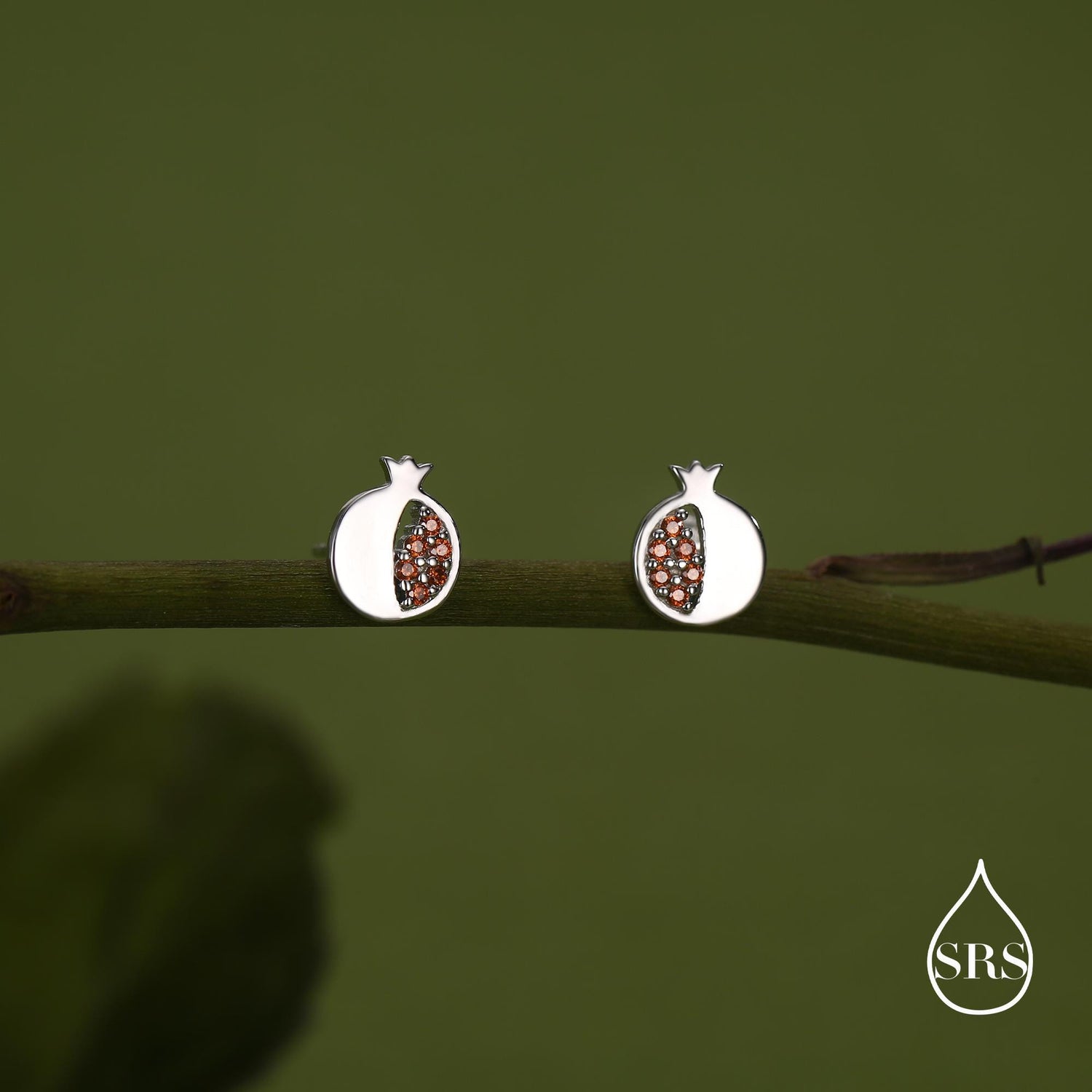 May include: A pair of silver-coloured stud earrings in the shape of pomegranates. The earrings have a white outer shell and are filled with small, reddish-brown stones. The earrings are displayed on a green branch against a green background.