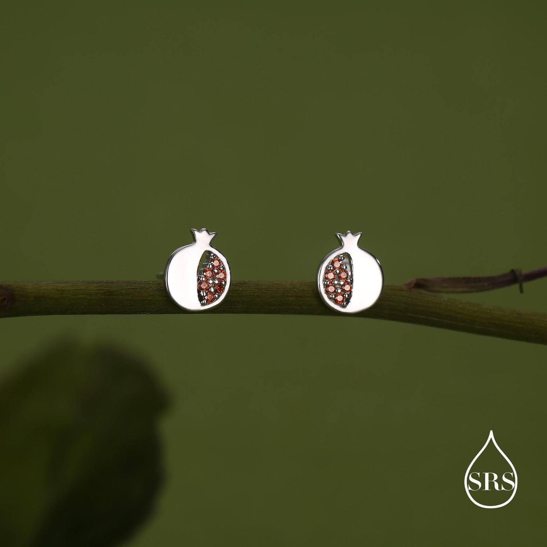 May include: A pair of silver-coloured stud earrings in the shape of pomegranates. The earrings have a white outer shell and are filled with small, reddish-brown stones. The earrings are displayed on a green branch against a green background.