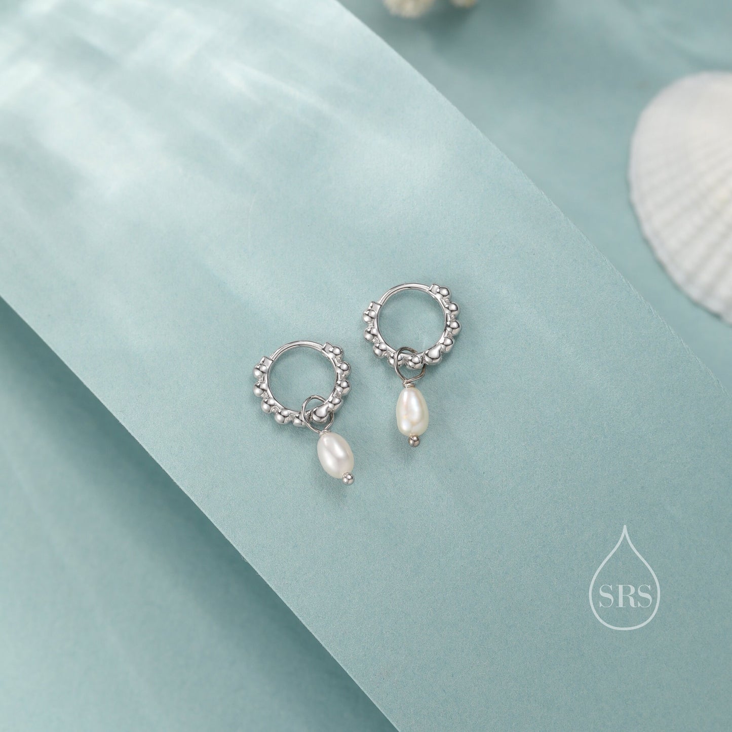 May include: A pair of silver hoop earrings with a small pearl dangling from each hoop. The earrings are on a light blue background.
