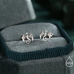 May include: Silver dragon stud earrings. Each earring showcases a detailed dragon design with outstretched wings and a curled tail. The earrings are presented in a teal velvet jewellery box, providing a striking contrast.