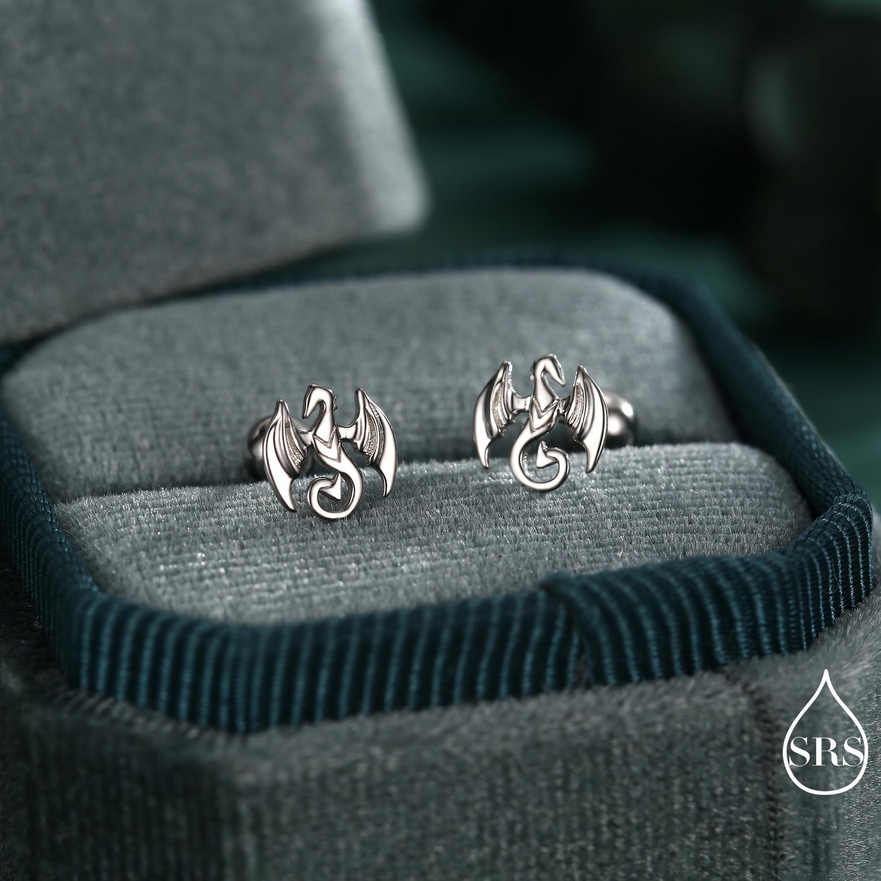 May include: Silver dragon stud earrings. Each earring showcases a detailed dragon design with outstretched wings and a curled tail. The earrings are presented in a teal velvet jewellery box, providing a striking contrast.