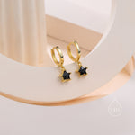 May include: Gold-coloured hoop earrings, each with a black star-shaped gemstone pendant. The earrings are presented on a light-toned surface, highlighting their design and colour.