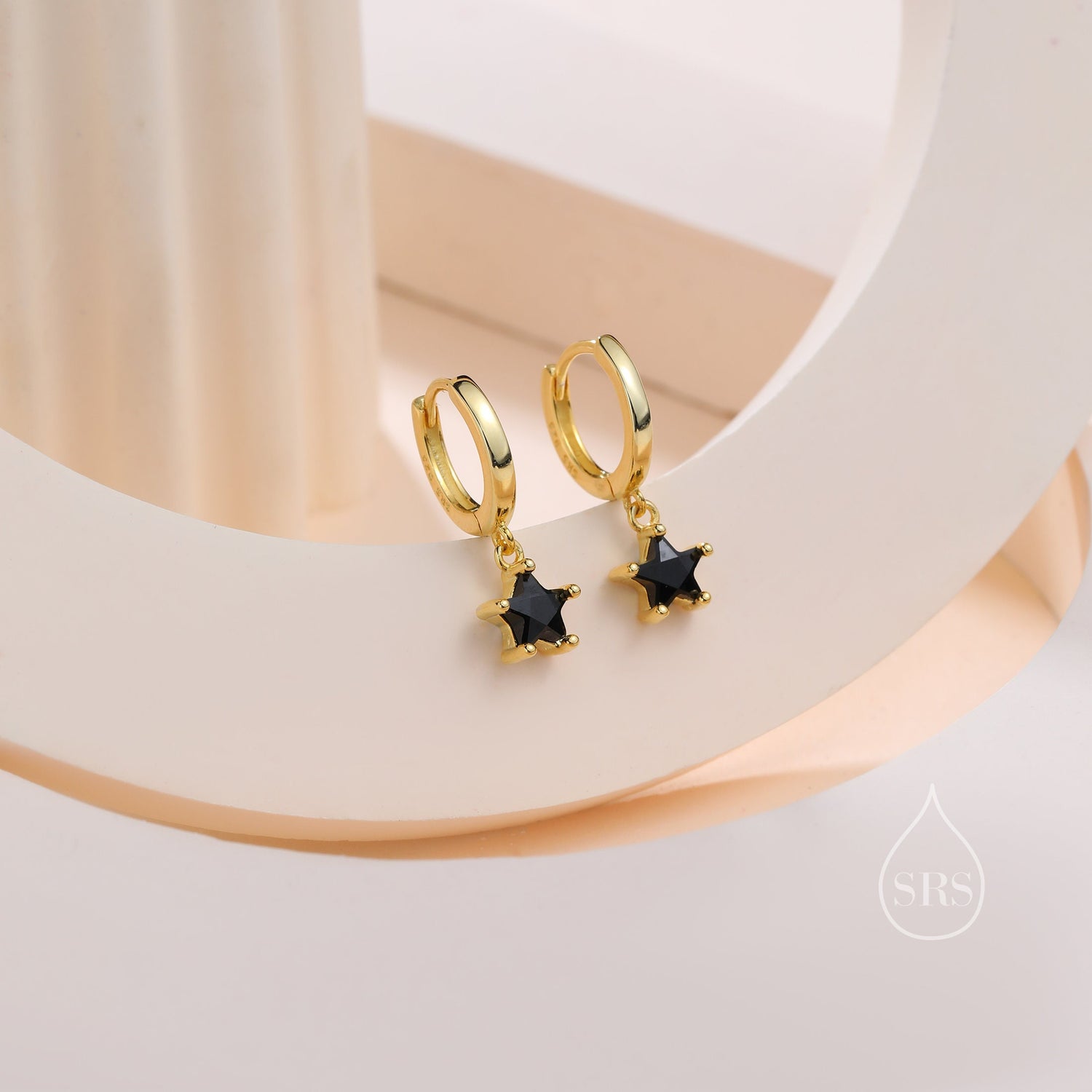 May include: Gold-coloured hoop earrings, each with a black star-shaped gemstone pendant. The earrings are presented on a light-toned surface, highlighting their design and colour.