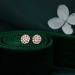 May include: A pair of small, round, rose gold earrings with a textured surface. The earrings are on a green velvet background.