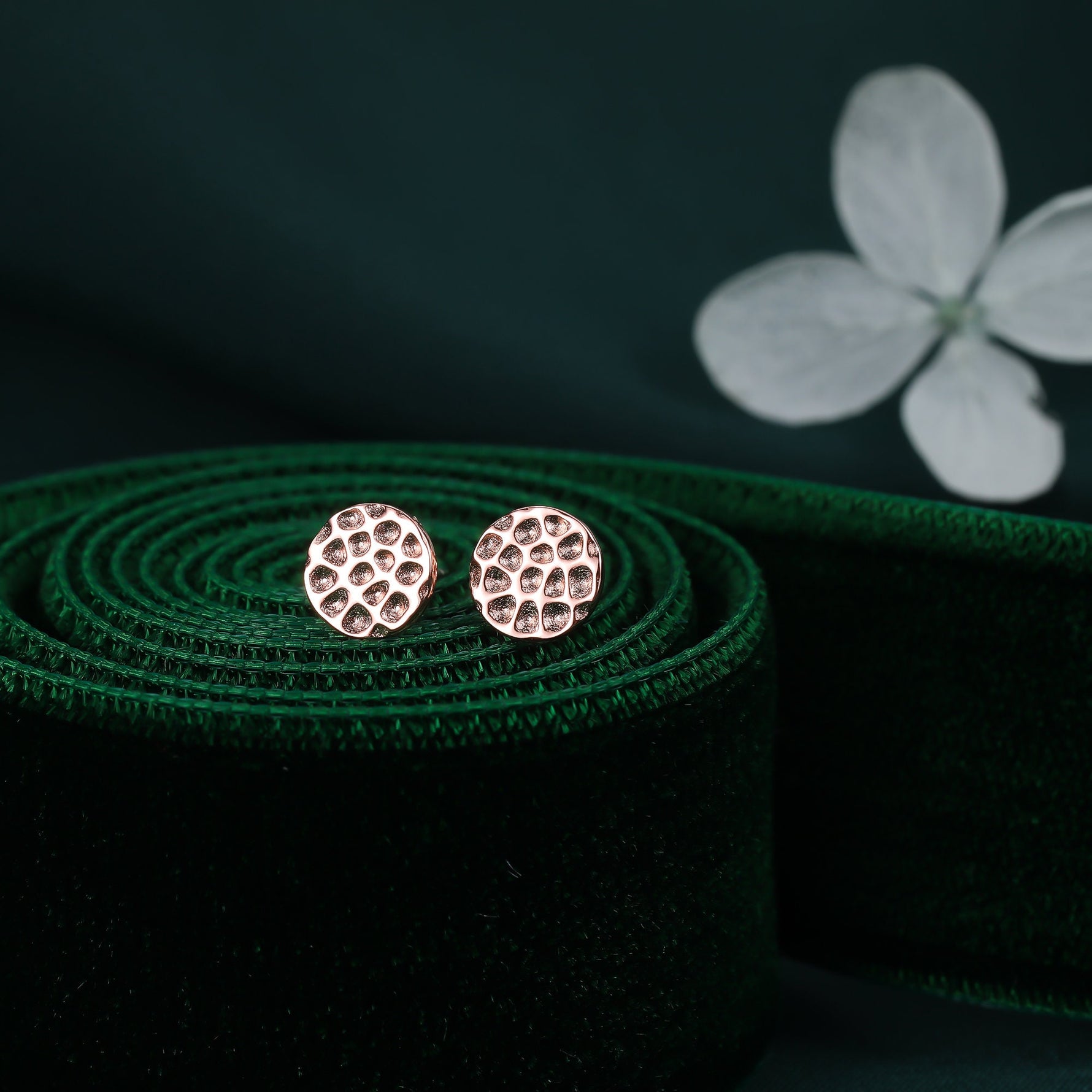 May include: A pair of small, round, rose gold earrings with a textured surface. The earrings are on a green velvet background.