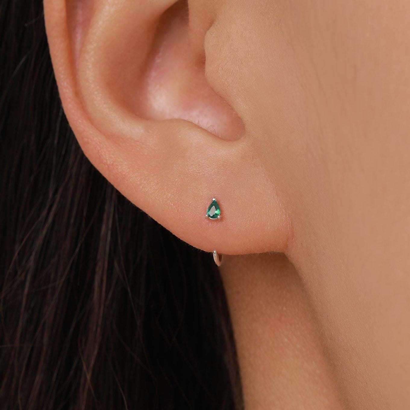 May include: A close-up of an ear with a small, teardrop-shaped, emerald-green stud earring. The earring is set in a silver or white metal. The ear has a small silver hoop earring in the lower lobe.